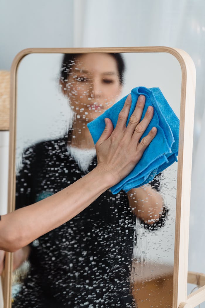 Home Close-up of a person wiping a mirror with a blue cloth, showing hands and reflection.